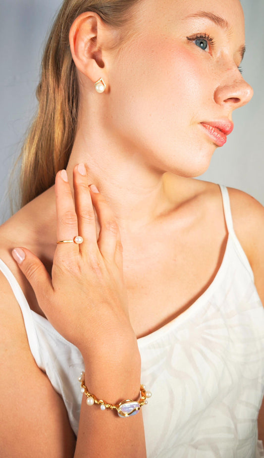 Woman wearing gold jewelry including earrings, ring, and bracelet on a neutral background