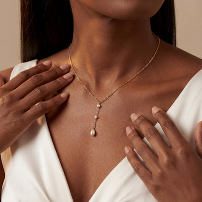 Woman wearing a delicate necklace with a pearl pendant, hands gently touching her shoulders.
