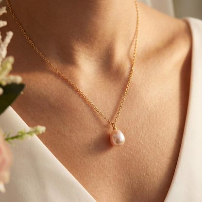 Gold necklace with a pink pearl pendant worn by a person, held by a blurred hand holding flowers.