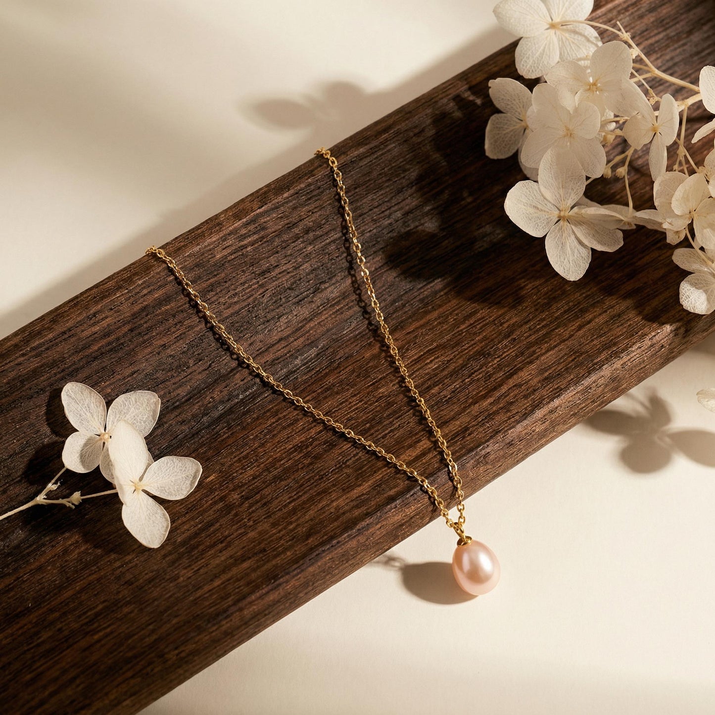 Gold necklace with a pink pearl pendant on a wooden surface with white flowers.