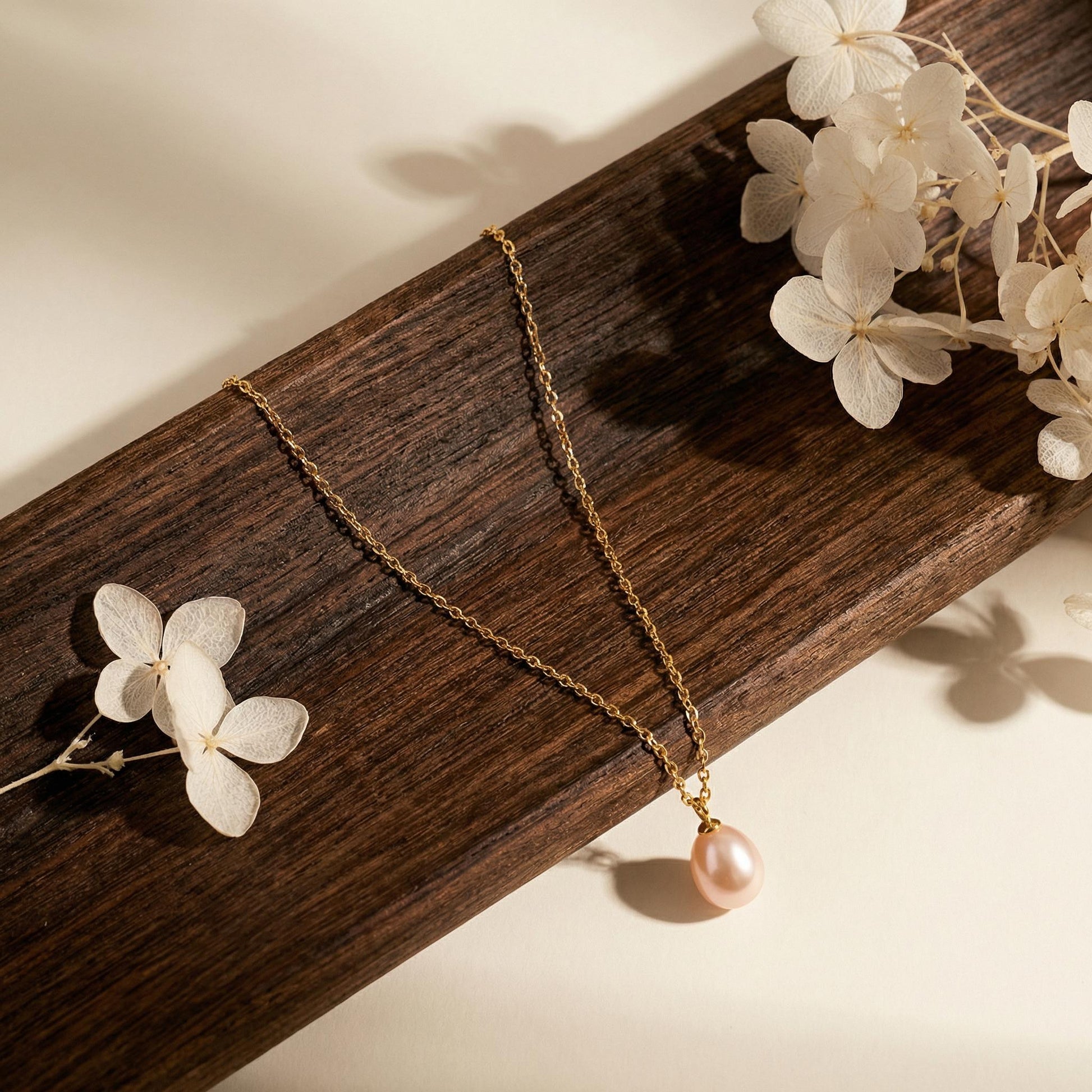 Gold necklace with a pink pearl pendant on a wooden surface with white flowers.